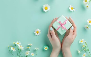 Mali poklon u mint boji sa roze mašnom koji drži devojka i cvetovi koji se nalaze oko njega.