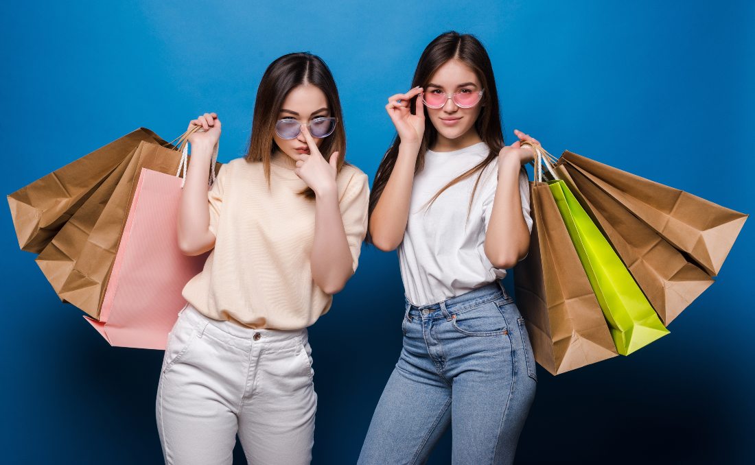 Dve devojke sa &scaron;oping kesama poziraju ispred plave pozadine.