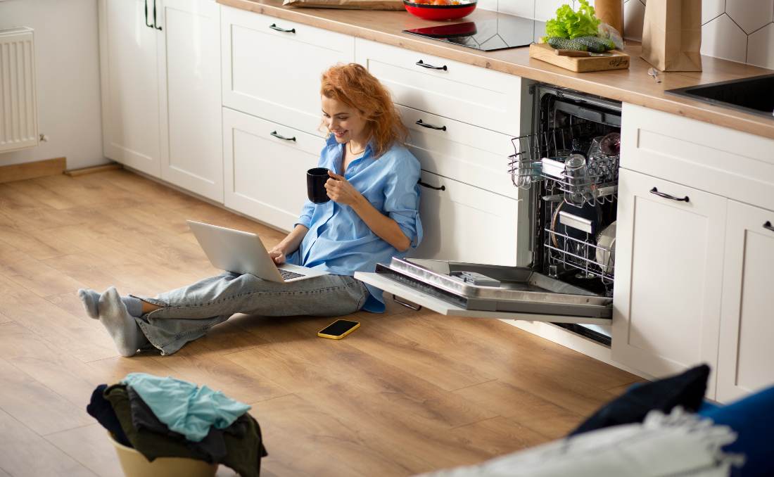 Žena stoji pored ma&scaron;ine za sudove u kuhinji i uživa u odmoru nakon pranja sudova.